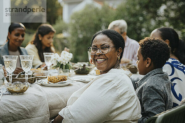 Portrait of happy senior woman with eyeglasses looking over shoulder while sitting with family members at lunch