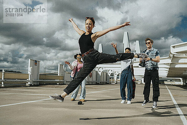 Young woman in casuals doing split while male and female friends applauding in parking lot under cloudy sky