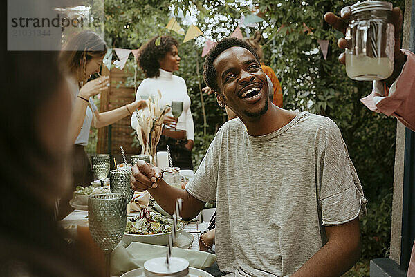 Happy young man eating salad and talking with friends in back yard party