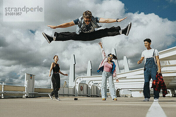 Young man doing split in mid-air while male and female friends standing in parking lot under cloudy sky