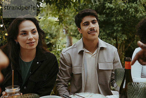 Young man and woman listening to friend while sitting at dining table in back yard