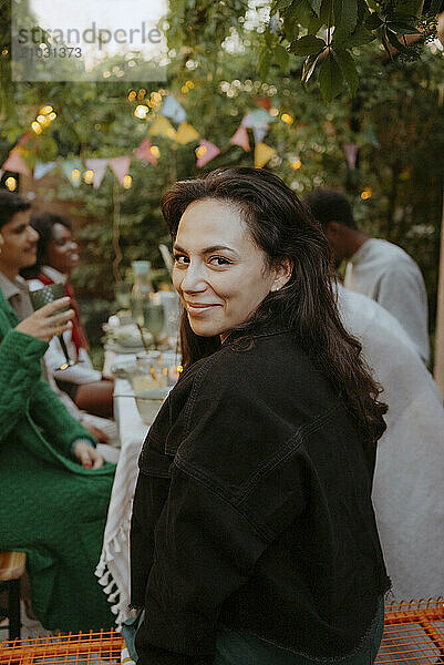 Portrait of smiling woman with long hair looking over shoulder while sitting with friends at dining table in back yard