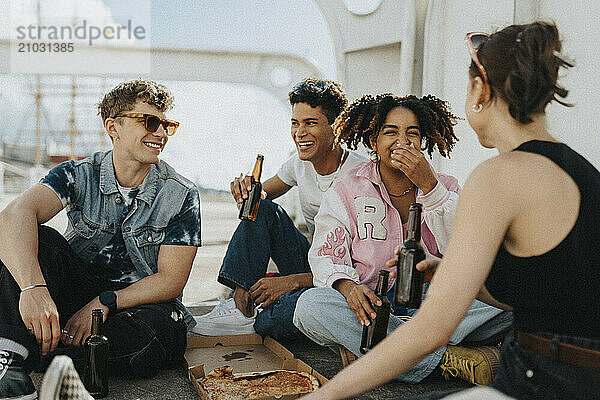 Happy young male and female friends having pizza with beer while sitting on ground at parking lot