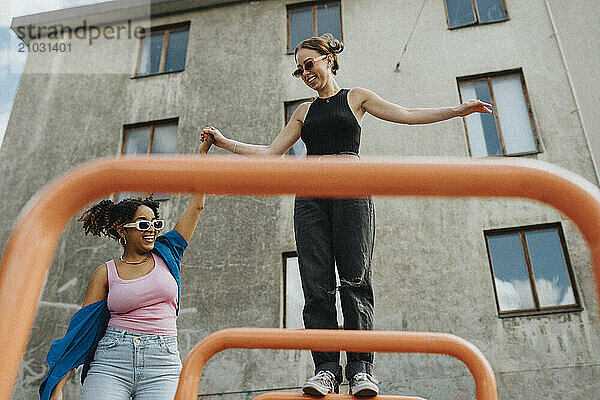 Happy young woman holding hands of female friend standing on metal bar in playground against building