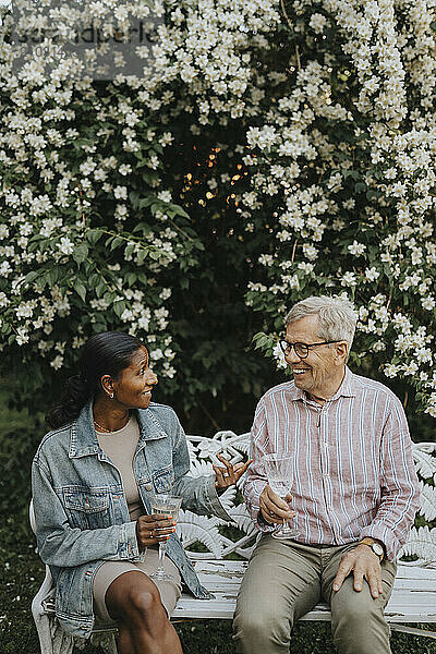 Mature woman and senior man holding glass while discussing and sitting on bench at garden party