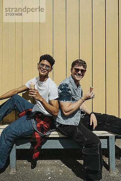 Portrait of fashionable young male friends holding ice cream while sitting on bench against corrugated wall