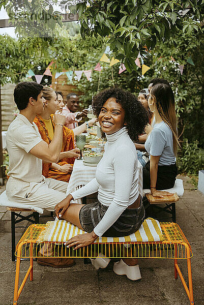 Side view portrait of happy young woman sitting on bench with friends at dining table during party