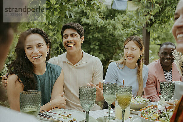 Happy male and female friends talking while sitting together at dining table in back yard