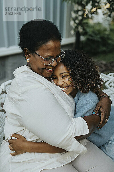 Happy grandmother embracing granddaughter while sitting on bench at back yard