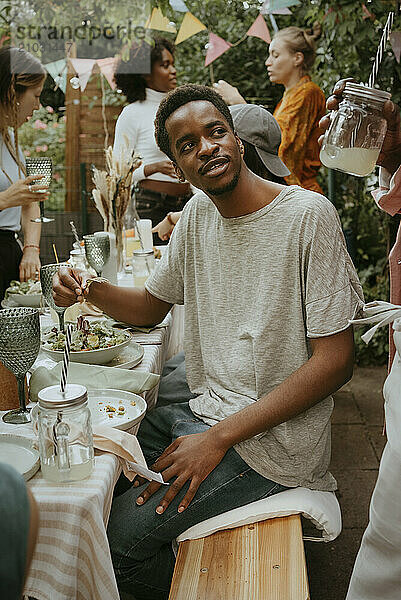 Young man eating salad and talking with friend while sitting on bench at dinner party in back yard