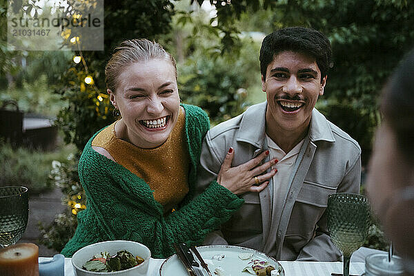 Cheerful male and female friends laughing while sitting at dining table in back yard