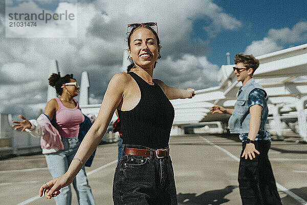 Portrait of beautiful young woman in casuals dancing with male and female friends in parking lot under cloudy sky