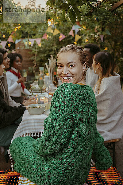 Portrait of happy woman in green sweater looking over shoulder while sitting with friends at dining table