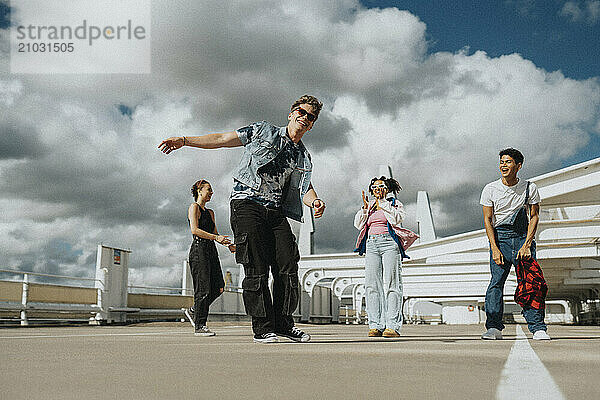 Low angle view of happy fashionable young man dancing with male and female friends standing in parking lot under cloudy