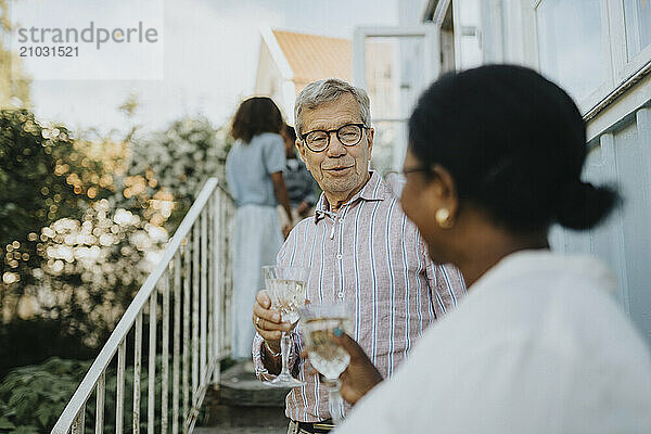 Senior man holding glass and talking with female family friend while standing on staircase outside house