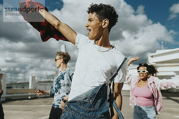 Cheerful hipster man in bib overalls dancing with male and female friends in parking lot