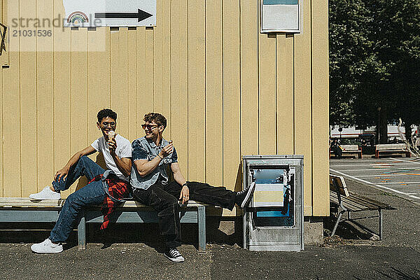 Carefree fashionable young male friends eating ice cream while sitting on bench against corrugated wall