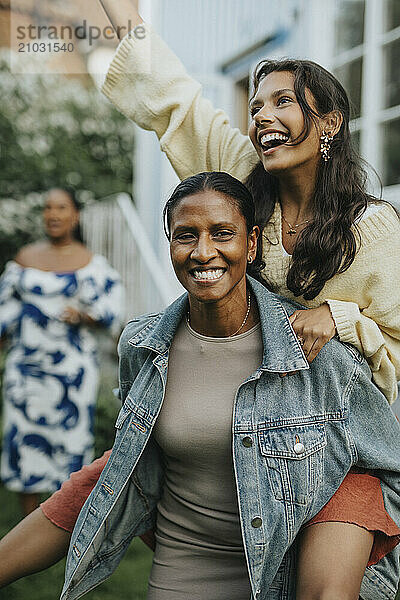 Happy mature woman piggybacking daughter at social gathering with family in back yard