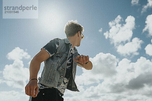 Low angle view of young man in denim jacket dancing under cloudy sky at sunny day