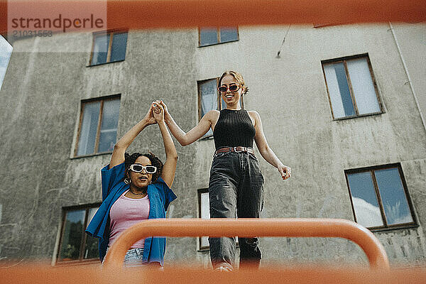 Young woman holding hands of female friend balancing on metal bar in playground against building