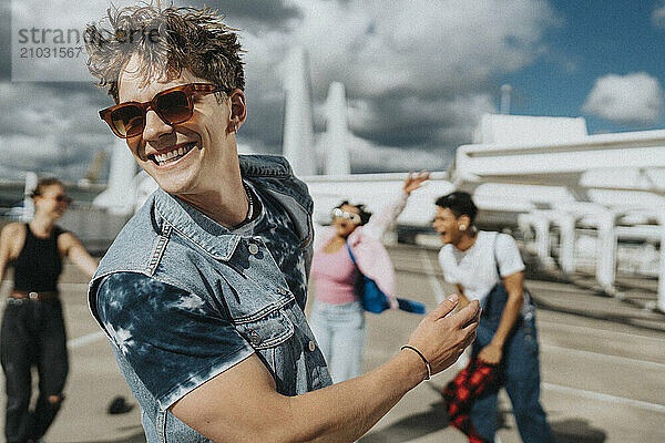 Cheerful young man in sunglasses with tattooed arm dancing with male and female friends in parking lot