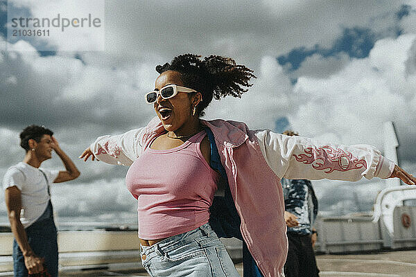 Carefree curly hair woman in pink letterman jacket with arms outstretched dancing at parking lot under cloudy sky