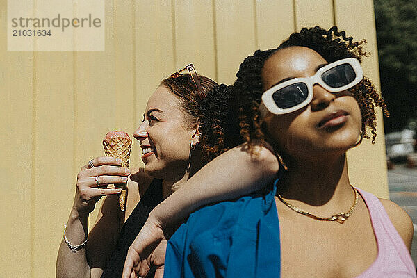 Cheerful young woman eating ice cream while leaning on fashionable female friend against corrugated wall