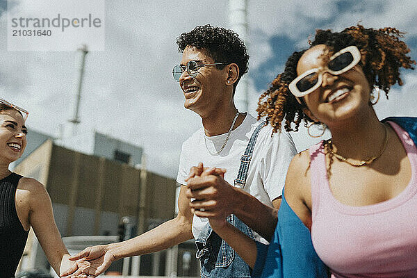 Happy fashionable young man holding hands and dancing with female friends at street