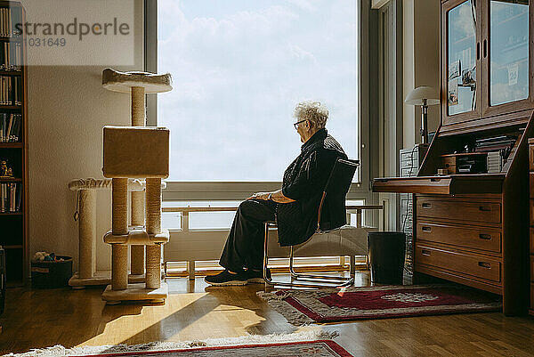 Full length of senior woman looking through window while sitting on chair in apartment