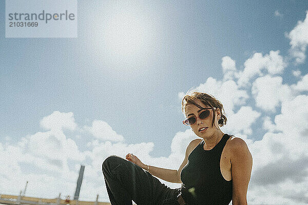 Portrait of young woman in sunglasses and black casuals posing under cloudy sky at sunny day