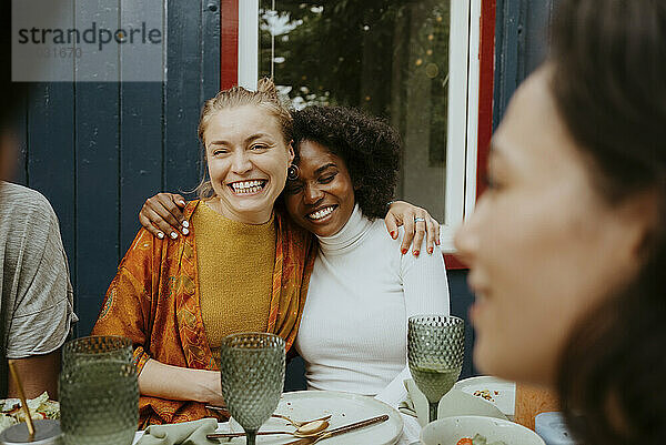 Happy multiracial female friends sitting with arms around while talking to friends at dinner party