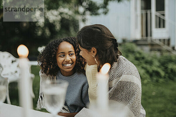 Happy teenage girl with arm around female friend sitting near table at family gathering