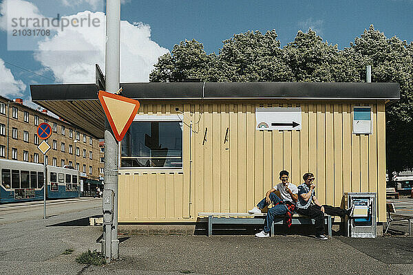 Fashionable young male friends eating ice cream while sitting on bench against corrugated wall in city