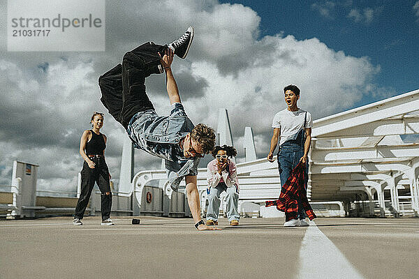 Acrobatic young man doing handstand while male and female friends cheering in parking lot under cloudy sky