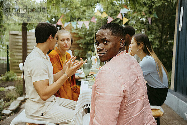 Portrait of smiling man looking over shoulder while sitting with friends celebrating at dinner party