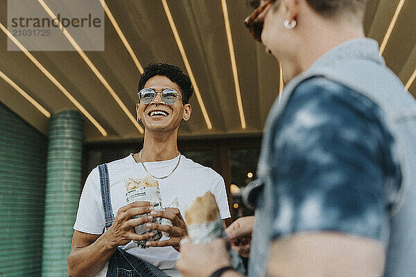 Happy young man in sunglasses holding wrap sandwich and standing with male friend