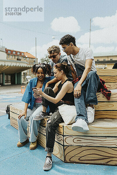 Curious young male and female friends sharing smart phone while sitting on steps at street