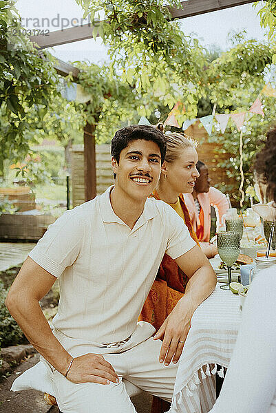 Smiling young man sitting with group of friends at dining table during dinner party in back yard