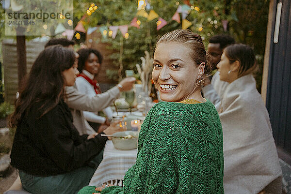 Portrait of cheerful young woman looking over shoulder while sitting with friends in back yard