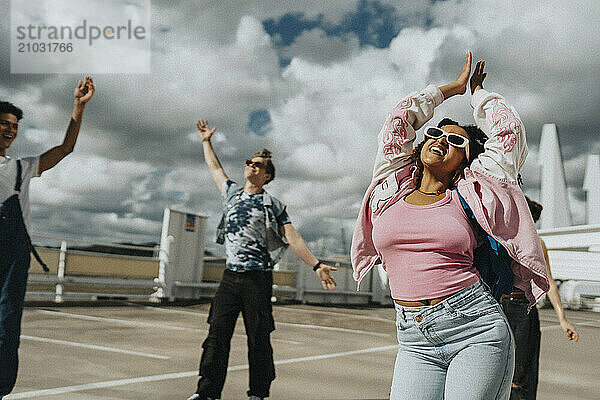 Cheerful young woman in letterman jacket clapping while dancing with friends at parking lot under cloudy sky