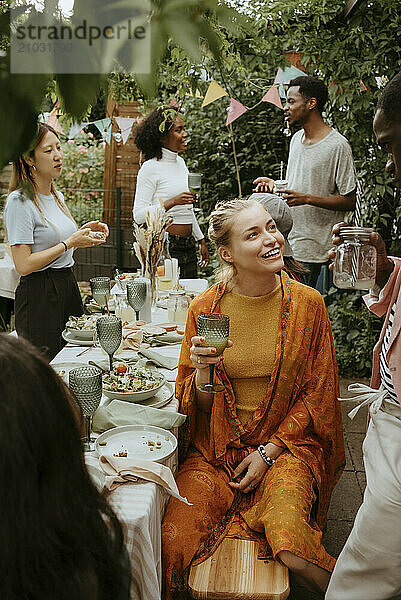 Smiling woman holding drink glass and talking with friend while sitting at dining table in party