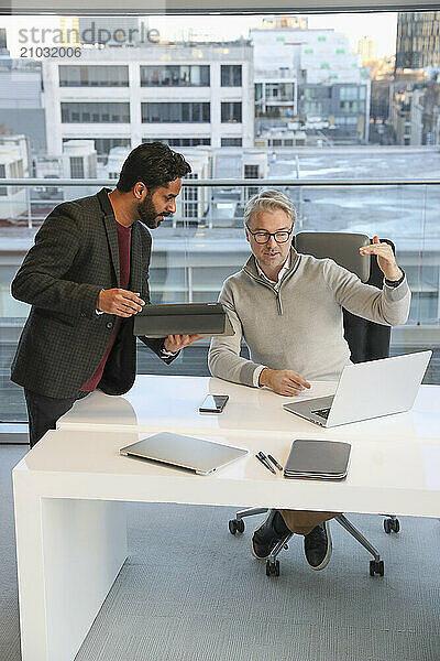 Businessmen talking in office; Two businessmen looking at laptop and talking in modern office at desk