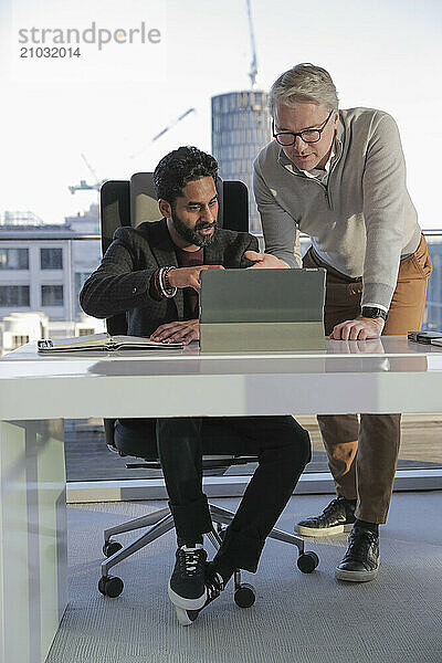 Businessmen talking in office; Two businessmen looking at digital tablet and talking in modern office at desk