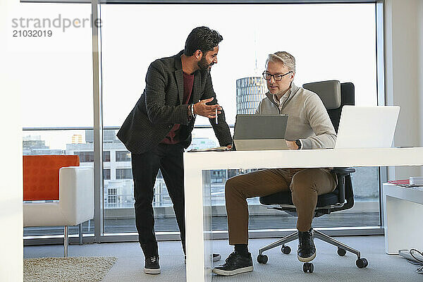 Businessmen talking in office; Two businessmen looking at digital tablet and talking in modern office at desk