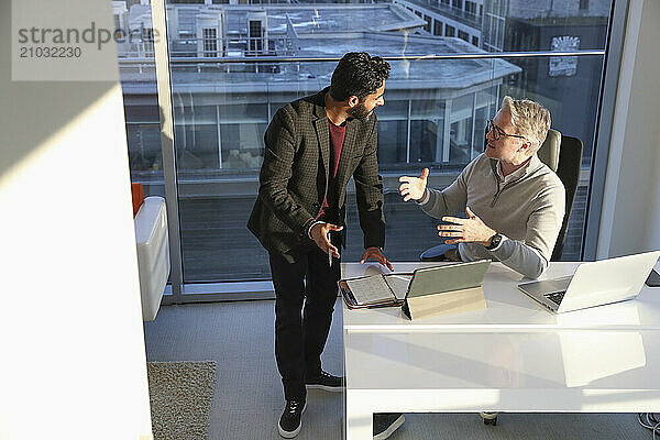 Businessmen talking in office; Two businessmen talking in modern office at desk with digital tablet and laptop