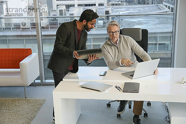 Businessmen talking in office; Two businessmen looking at laptop and talking in modern office at desk