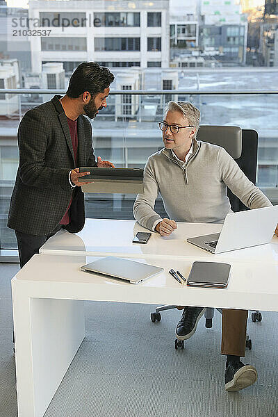 Businessmen talking in office; Two businessmen looking at digital tablet and talking in modern office at desk