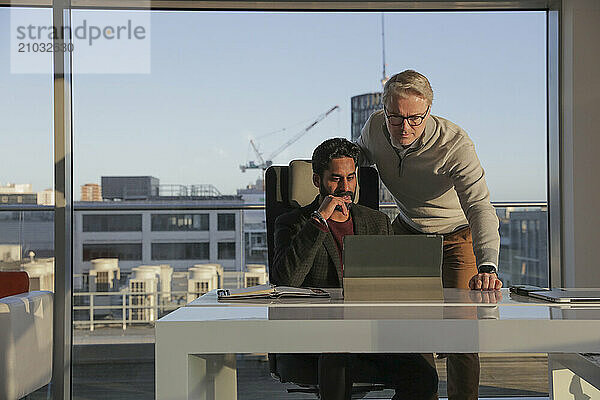 Businessmen talking in office; Two businessmen looking at digital tablet and talking in modern office at desk