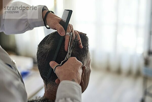 A hairdresser in a white shirt is cutting a man's hair with a pair of scissors and a comb at home