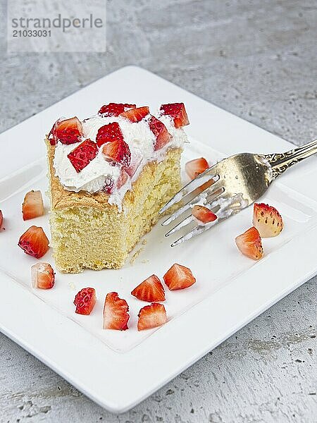 A close up photo of a slice of homemade sponge cake topped with whipped cream and cut up srtawberries and a corner cut out